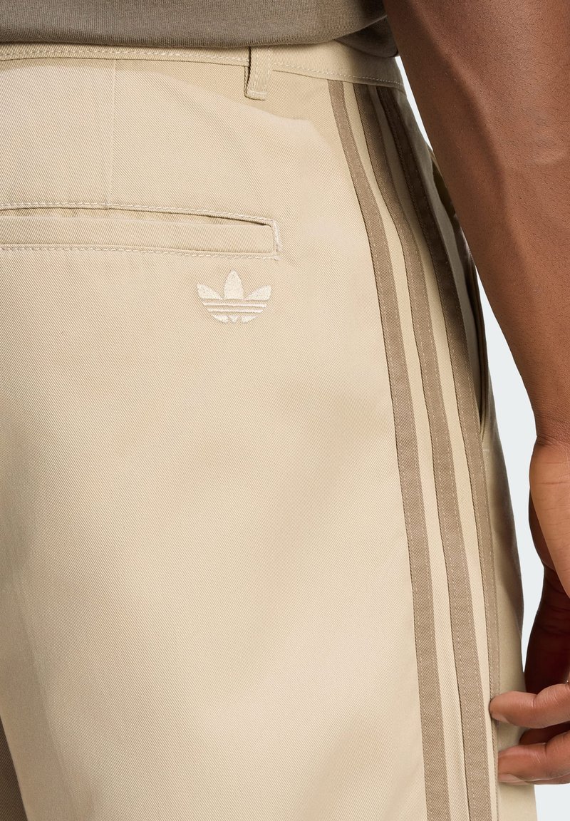 Beige pants with three vertical stripes on the side and a small Adidas logo near the back pocket, worn by a person with dark skin.