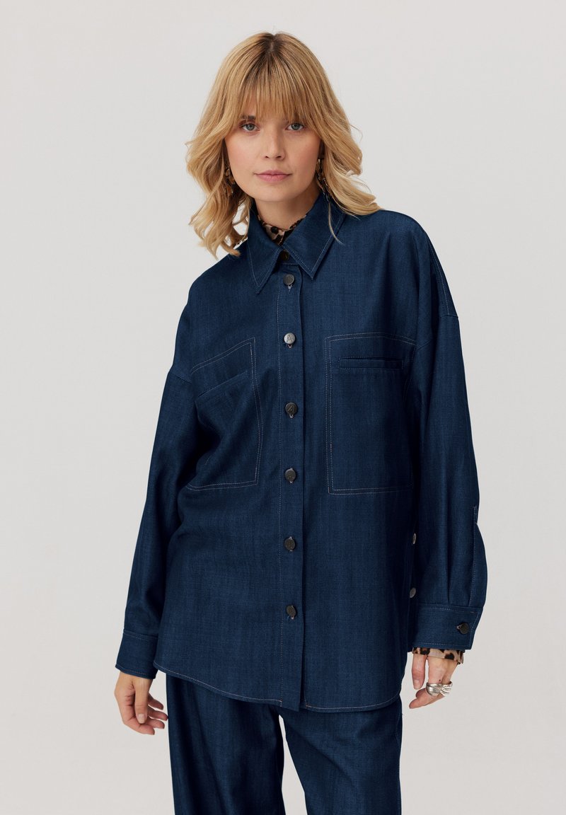 Blondynka w ciemnoniebieskiej, obszernej koszuli zapinanej na guziki oraz dopasowanych spodniach, stojąca na tle jasnym.