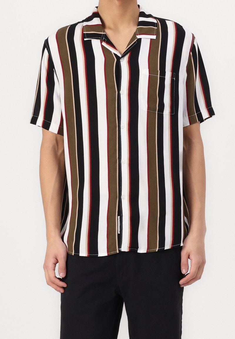 Personne portant une chemise à manches courtes boutonnée avec des rayures verticales noires, blanches, marron et rouges, et un pantalon noir, debout.