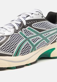 Sportovní bota s bílou síťovinou, šedými akcenty, zelenými dekorativními liniemi a polstrovanou béžovou mezipodešví. Texturovaná černá gumová podrážka.