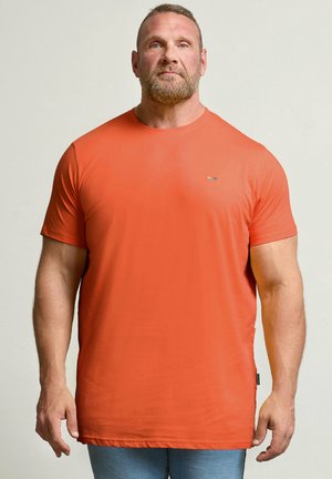 Man met baard die een simpele oranje korte mouw T-shirt en blauwe spijkerbroek draagt, staand tegen een lichte neutrale achtergrond.