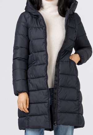 Femme portant un long manteau matelassé noir avec capuche, un pull à col roulé blanc en dessous et un jean bleu, une main dans la poche du manteau.