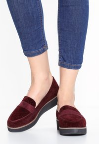 Mocassins en suède bordeaux avec une pointe arrondie, des détails en forme de vagues sur les bords, une semelle plate noire et un dessus texturé. Jeans visibles au-dessus des chaussures.