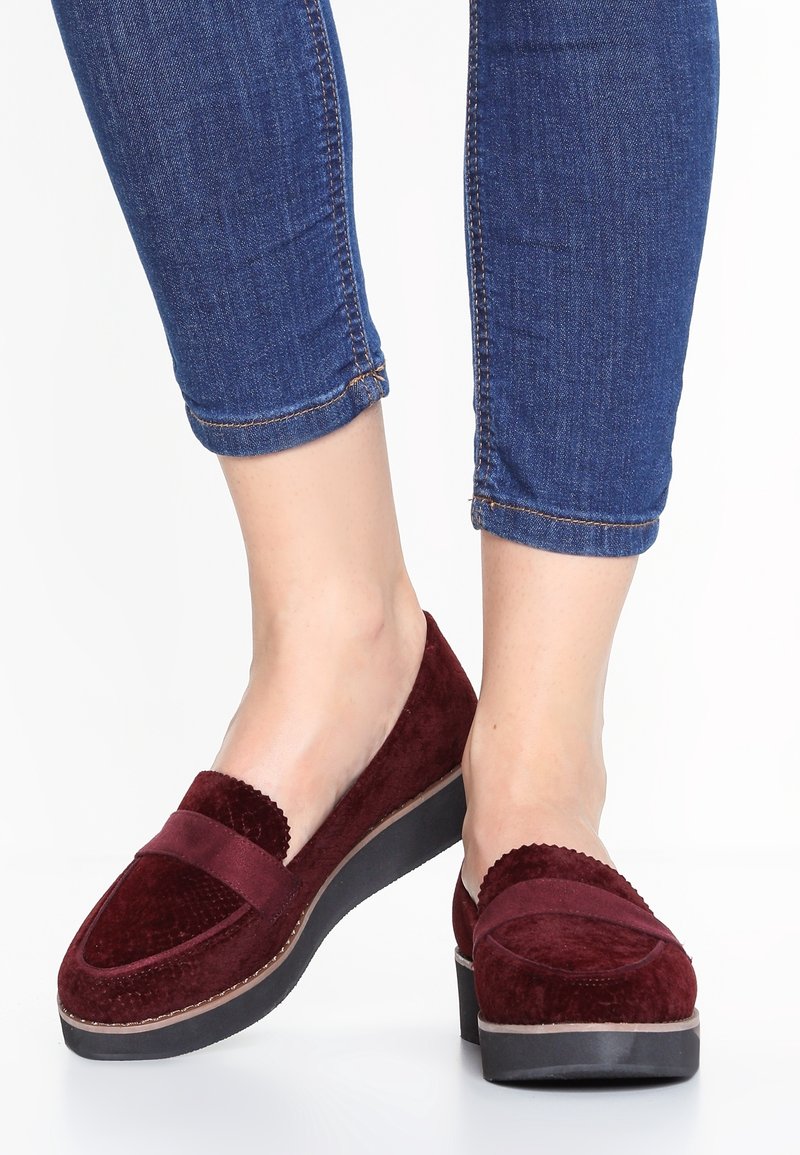 Mocassins en suède bordeaux avec une pointe arrondie, des détails en forme de vagues sur les bords, une semelle plate noire et un dessus texturé. Jeans visibles au-dessus des chaussures.