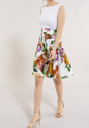 Haut blanc sans manches avec une jupe plissée à fleurs ornée de roses multicolores et de feuilles sur fond blanc. Talons dorés à lanières.