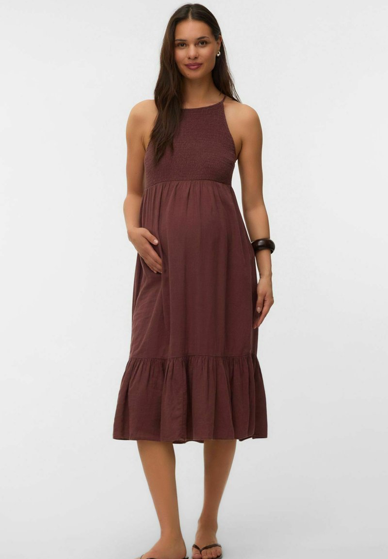 Femme aux longs cheveux foncés portant une robe midi marron sans manches, debout avec une main sur l'abdomen et un bracelet au poignet.