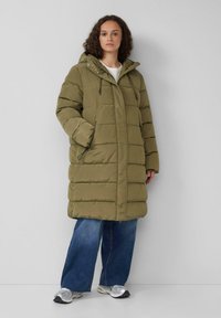 Olivgrüner gepolsterter Mantel mit Kapuze, mit gestepptem Muster und zwei seitlichen Taschen. Kombiniert mit lockeren blauen Jeans und silbernen Sneakern.