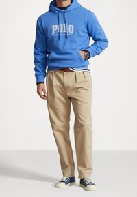 Man i blå Polo Ralph Lauren hoodie, beige byxor med brunt bälte, blå sneakers och en armbandsklocka, stående med en hand i fickan.