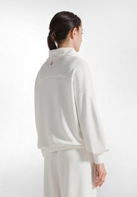 Suéter blanco con cuello alto, mangas amplias y dobladillo elástico. Presenta una pequeña etiqueta rosa en la parte posterior. Textura de tela suave y lisa.