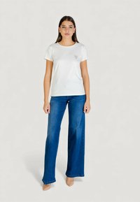 Biały bawełniany t-shirt z krótkimi rękawami i małym nadrukowanym logo po lewej stronie, w połączeniu z niebieskimi rozkloszowanymi dżinsami i beżowymi szpilkami.