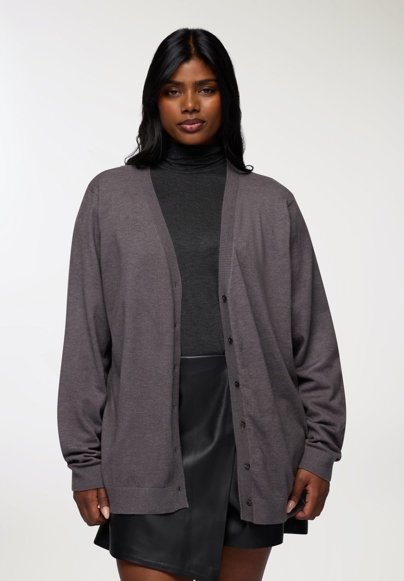 Cardigan grigio realizzato in materiale morbido, con vestibilità ampia, maniche lunghe, scollatura a v e chiusure a bottoni sulla parte anteriore. Abbinato a un top scuro.