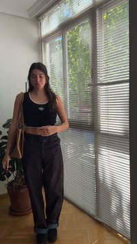 Νεαρή γυναίκα με μαύρο crop top και παντελόνι στέκεται κοντά σε παράθυρο με περσίδες κρατώντας γυαλιά ηλίου και καφέ τσάντα μέσα στο δωμάτιο.