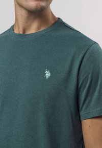 Majica tirkizne boje od mekanog pamuka, s okruglim izrezom i kratkim rukavima. Mali logotip svijetloplave boje na lijevoj strani prsa.