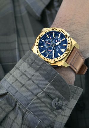 Reloj de metal dorado con una esfera azul, tres subesferas, marcadores de hora blancos y correa de cuero marrón, llevado sobre una camisa de cuadros grises.