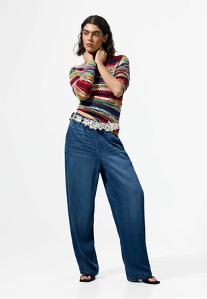 Junge Person, die einen bunten, gestreiften Pullover, weit geschnittene blaue Jeans, einen floral gemusterten Gürtel und schwarze Sandalen trägt, steht mit den Händen nahe am Hals.