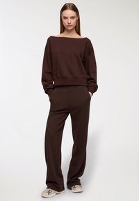 Felpa in cotone marrone con scollo ampio e maniche lunghe, abbinata a pantaloni marroni coordinati. Il modello indossa sneaker beige e grigie.