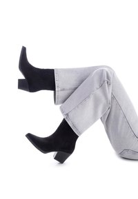 Botas de ante negras hasta el tobillo con un tacón cuadrado, que presentan una punta afilada y un diseño bordado. Combinadas con jeans rectos de color gris claro.