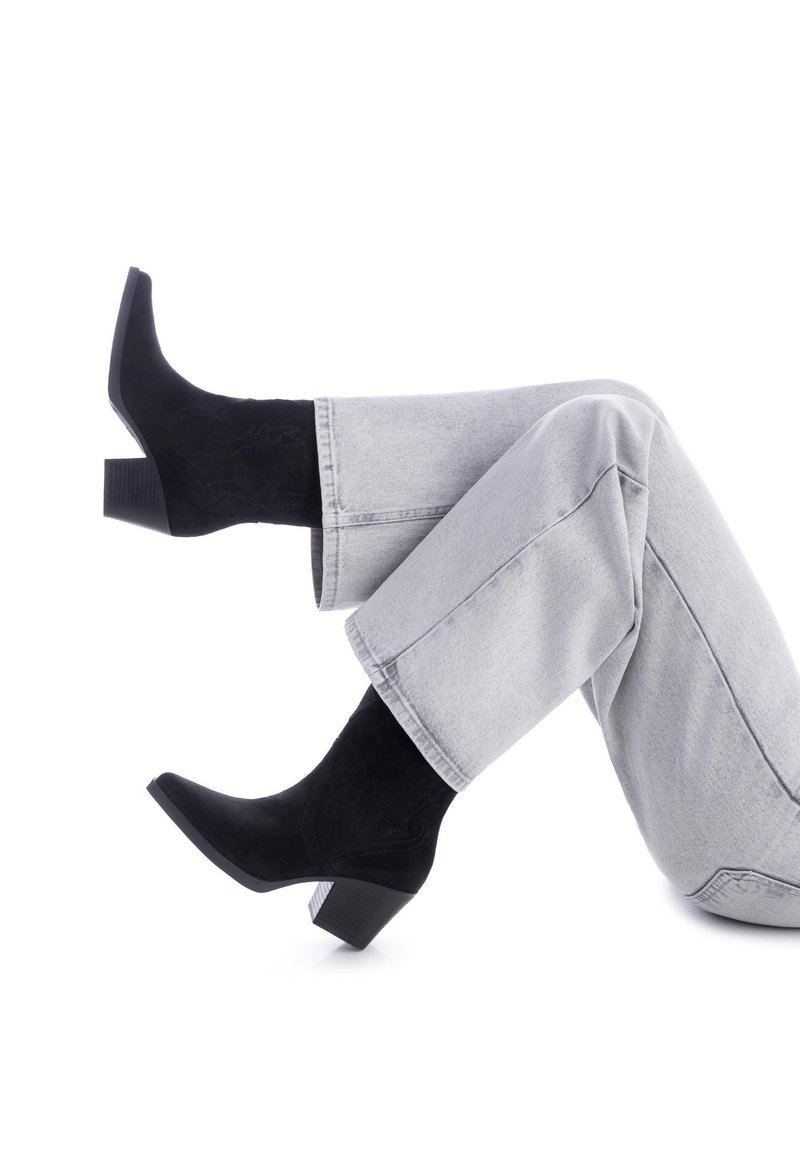 Botas de ante negras hasta el tobillo con un tacón cuadrado, que presentan una punta afilada y un diseño bordado. Combinadas con jeans rectos de color gris claro.