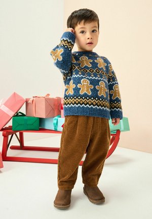 Blauer Strickpullover mit Lebkuchen- und Diamantmustern, kombiniert mit braunen Cordhosen und braunen Stiefeln, vor einem Hintergrund mit Feiertagsgeschenken.