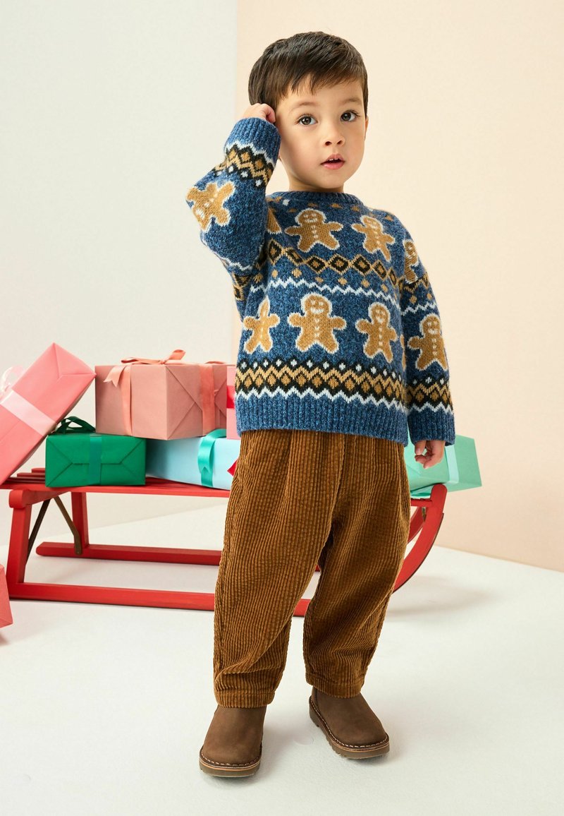 Blauer Strickpullover mit Lebkuchen- und Diamantmustern, kombiniert mit braunen Cordhosen und braunen Stiefeln, vor einem Hintergrund mit Feiertagsgeschenken.