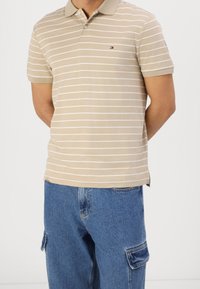 Beige gestreepte polo met korte mouwen en een kraag. Kenmerkt zich door witte horizontale strepen en een klein logo op de linkerborst. Gecombineerd met blauwe cargobroek.