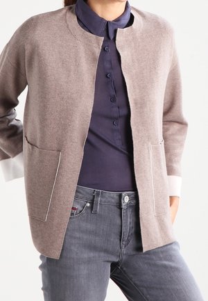 Cardigan marron clair en matériau doux, avec un col rond, une ouverture devant, deux poches et des poignets blancs. Porté sur une chemise foncée.