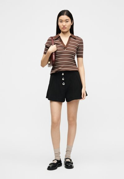 Jeune femme portant un polo rayé marron, un short taille haute noir à boutons, des chaussettes beiges, des mocassins noirs, et tenant un sac à bandoulière bordeaux.