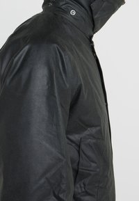 Černá nepromokavá bunda s vysokým límcem, zapínáním na druky a bočními kapsami, s hladkou, mírně lesklou texturou a strukturovaným designem.