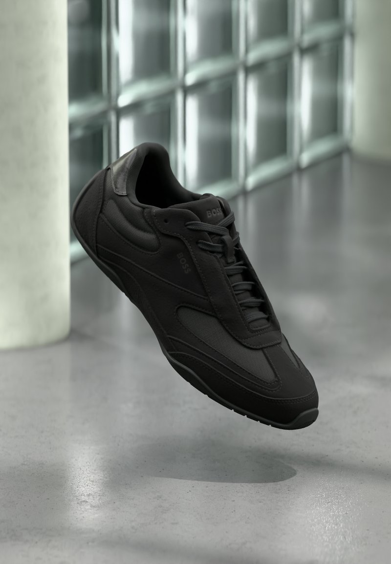 Czarny sneaker BOSS z teksturowanymi i gładkimi panelami, sznurowadła zawiązane, zawieszony nad polerowaną podłogą obok matowych szklanych bloków i białej kolumny.