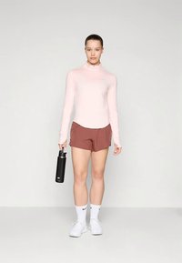 Hellrosa langärmliges Oberteil mit einem halben Reißverschluss, kombiniert mit braunen Sportshorts. Schwarze Wasserflasche in einer Hand gehalten. Weiße Turnschuhe und Socken.