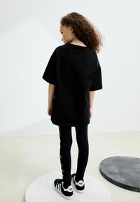 Crna oversized majica, crne tajice s bijelim bočnim naglascima i crne tenisice s bijelim prugama; dijete stoji i gleda prema van.
