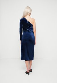 Μίντι φόρεμα από βελούδο με έναν μακρύ μανίκι, σκούρο μπλε χρώμα, κομψή εφαρμογή και μαλακή υφή. Μήκος μέχρι το γόνατο. Μαύρα ψηλοτάκουνα παπούτσια.