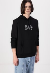 Hoodie noir en mélange de coton, avec une poche frontale et un logo "GAP" brodé en blanc. Manches longues et capuche avec cordon de serrage réglable.