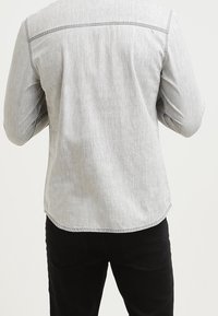 Chemise gris clair à ourlet arrondi, avec une texture subtile et des coutures contrastées le long des épaules, associée à un pantalon noir.