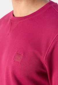 Gros plan d'une personne portant un sweat-shirt rose vif avec un petit logo carré embossé indiquant "BOSS" sur la poitrine.