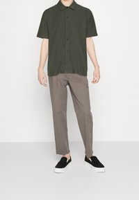 Uomo che indossa una camicia a maniche corte di colore verde oliva scuro, pantaloni color taupe e scarpe slip-on nere, in piedi davanti a uno sfondo bianco.