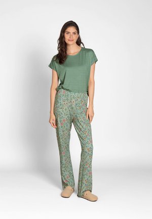 Woman standing wearing a green striped short-sleeve top, floral patterned green pants, and beige slippers against a plain light background.