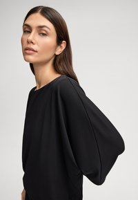Blouse noire avec des manches oversized et drapées, col rond, fabriquée dans un tissu lisse avec un léger éclat, peu de détails.