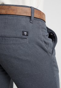 Acercamiento a unos pantalones grises texturizados con un cinturón de cuero marrón y una pequeña etiqueta con logo negro cerca del bolsillo trasero.