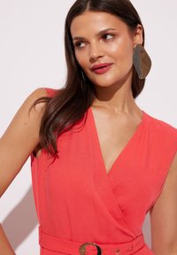 Koronki sukienka w kolorze koralowym z dekoltem w kształcie litery V i krótkimi rękawami. Posiada złotą okrągłą klamrę w talii. Modelka nosi duże metaliczne kolczyki w kształcie liści.