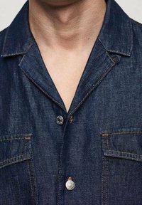 Ciemnoniebieska koszula jeansowa z ostrym kołnierzem, dwoma kieszeniami na piersi i srebrnymi metalowymi guzikami. Charakteryzuje się przeszyciami dekoracyjnymi i gładką fakturą.