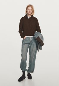 Donna con capelli biondi corti che indossa un maglione marrone a trecce, jeans larghi di un blu chiaro, calze scure e tiene una giacca di denim.