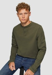 Oliwkowa, długosleeve'owa koszula henley z okrągłym dekoltem, plisą na dwa guziki i miękkiej bawełnianej tkaniny. W zestawie z niebieskimi dżinsami.