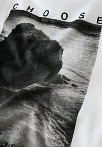 Witte T-shirt met een zwart-wit grafiek van een rotsachtig strandscène met de tekst "KIES EEN GELUKKIG LEVEN" bovenop de afbeelding. Gladde textuur.