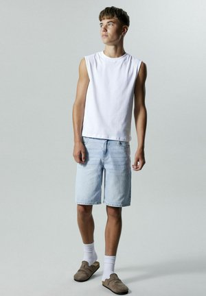 Biała, bezrękawnikowa koszula bawełniana w zestawie z jasnoniebieskimi szortami dżinsowymi, beżowymi butami na slip-on oraz białymi skarpetkami. Prosty fason i luźny krój.