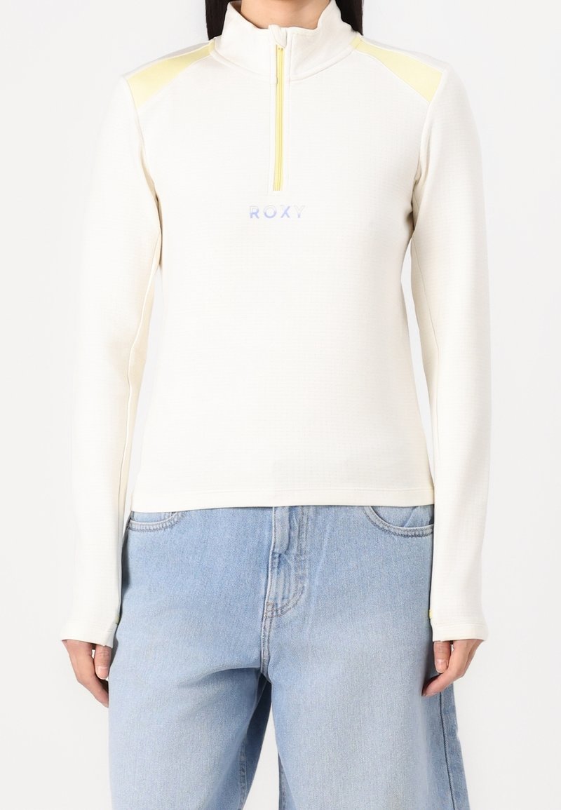 Femme portant un pull crème à manches longues avec une fermeture éclair au quart, bandes jaunes sur les épaules et un jean bleu clair, debout sur un fond blanc.