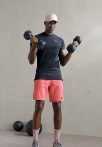 Svart kortermet skjorte, rosa shorts, holder svarte sekskantede manualer. Grå sneakers og lyse rosa caps. Minimalistisk bakgrunn.