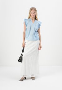Blusa sin mangas de color azul claro con bordes con volantes, combinada con una falda maxi blanca de encaje. La modelo sostiene un bolso negro y lleva tacones beige.