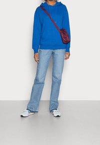 Felpa blu con cappuccio e tasca frontale, jeans a zampa d'elefante blu chiaro e sneaker bianche con accenti multicolori. Borsa a tracolla bordeaux.