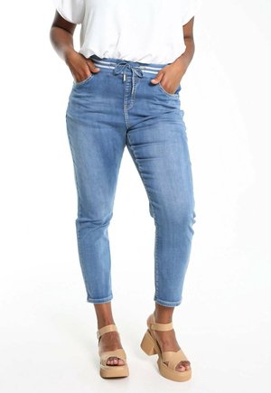 Personne portant un pantalon jogger en denim bleu clair avec une taille à cordon et des sandales à plateforme beiges, mains dans les poches.